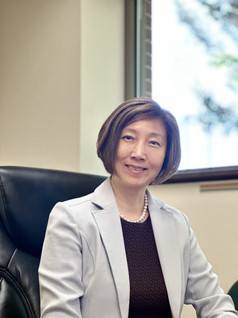 Professional woman in a light gray blazer with pearl necklace, seated in an office chair near a window.