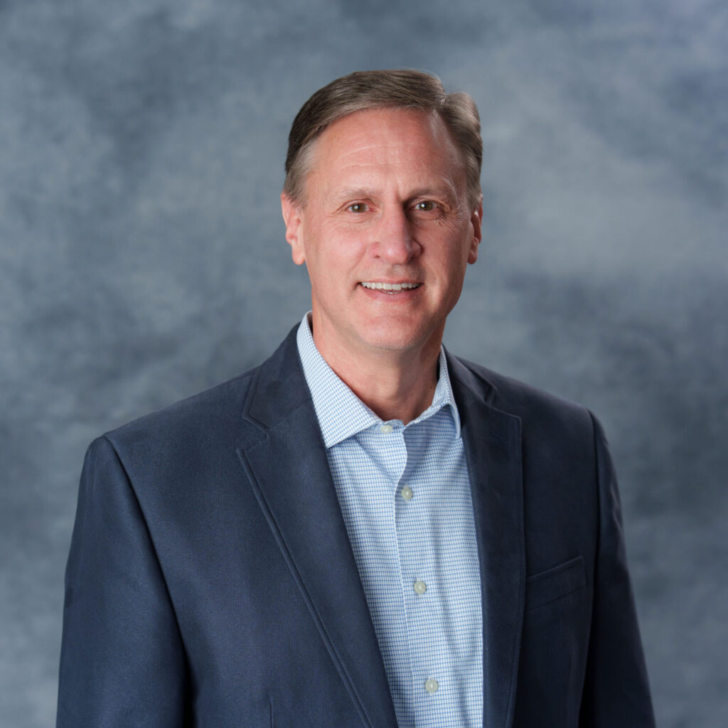 Portrait of a smiling man in a dark blazer and light blue checkered shirt, shown from the chest up against a blue-gray backdrop.